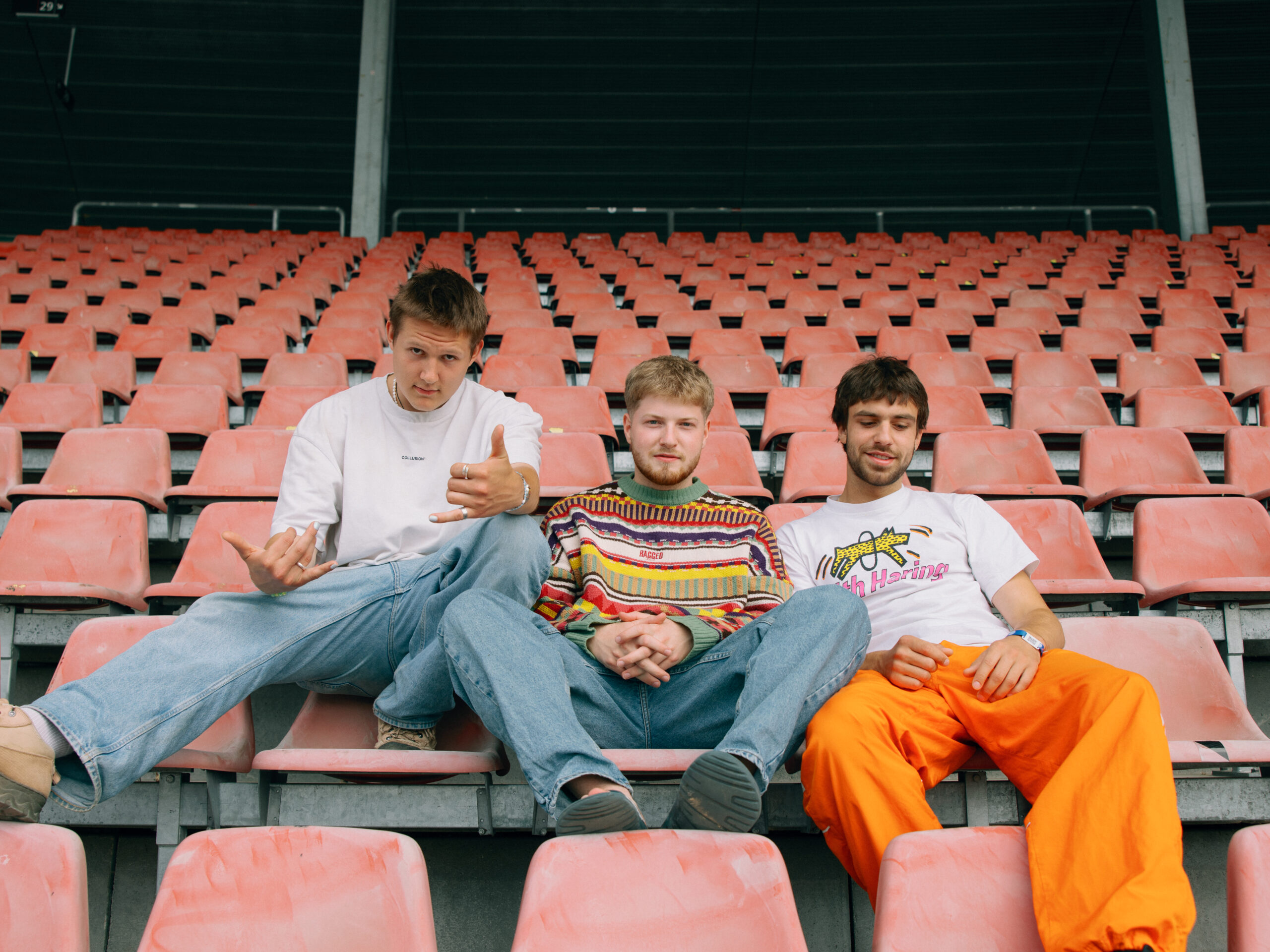 Drei junge Männer, die zusammen die Band 01099 bilden sitzen auf dem Bild nebeneinander auf den orangen Stühlen der Tribüne eines Fußballstadions. Die drei sitzen lässig eng neben einander und posen in die Kamera.